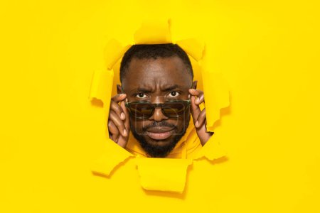 Foto de Hombre afroamericano serio mirando a la cámara a través de gafas de sol, notando algo, rompiendo a través de fondo de papel amarillo, espacio libre - Imagen libre de derechos