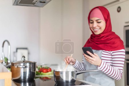 Téléchargez les photos : Belle jeune femme musulmane portant hijab cuisine à la maison, lecture blog culinaire sur Internet, en utilisant un téléphone portable moderne tout en faisant un dîner sain pour sa famille, copier l'espace. concept blog alimentaire - en image libre de droit