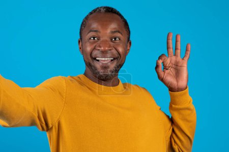 Photo for Closeup of happy handsome middle aged black man in yellow taking selfie on blue studio background, cheerfully smiling at camera, showing okay gesture, posting photo on social media, copy space - Royalty Free Image
