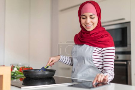 Foto de Hermosa joven musulmana vistiendo hijab cocinar en casa, leyendo blog culinario en Internet, utilizando tableta digital mientras hace una cena saludable para su familia, espacio de copia. Concepto blog de alimentos - Imagen libre de derechos