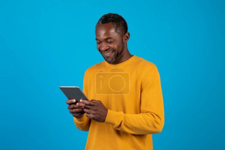 Téléchargez les photos : Studio shot of happy adult Afro-Américain en tenue décontractée en utilisant une tablette numérique flambant neuve isolée sur fond bleu, jeu sur Internet, en utilisant une belle application mobile divertissante, espace de copie - en image libre de droit