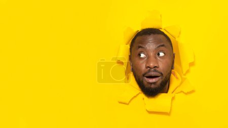 Foto de Omg. Hombre negro sorprendido mirando a un lado el espacio libre a través de un agujero en papel amarillo roto, posando con la boca abierta en el estudio, panorama. Sorprendida oferta - Imagen libre de derechos
