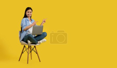 Glad cute millennial korean lady student in casual show fingers at empty space, sit on chair with laptop, isolated on yellow background, studio. Study, work, blogger advice, huge sale, ad and offer