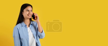 Photo for Glad cute millennial korean female in casual calling by smartphone, talking with friend, look at copy space, isolated on yellow background, panorama. Gossip and good news, communication, ad and offer - Royalty Free Image