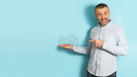 Photo for Excited caucasian mature man pointing finger at open palm, posing on blue background, panorama with free space. Cheerful male advertising something, showing invisible object - Royalty Free Image