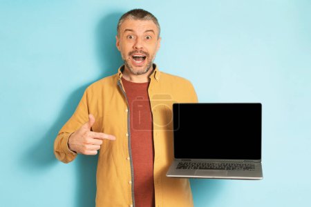 Téléchargez les photos : Heureux homme d'âge moyen pointant vers l'écran d'ordinateur portable vide avec maquette pour la conception de site Web sur fond bleu studio. Homme émotionnel montrant ordinateur avec espace pour votre annonce - en image libre de droit