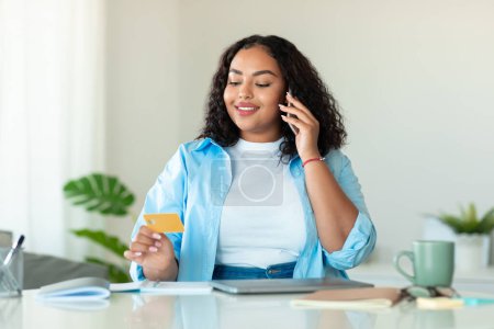 Foto de Concepto de banca online. Mujer alegre cuerpo negro positivo hablando en el teléfono celular y la celebración de la tarjeta de crédito, sentado en la mesa en casa. Afro-americana dama haciendo pagos en internet - Imagen libre de derechos