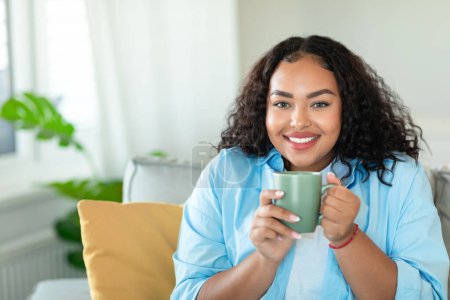 Foto de Ocio doméstico. Happy black plus size lady bebiendo café en casa, descansando en un cómodo sofá en la acogedora sala de estar, sosteniendo la taza con bebida caliente y sonriendo - Imagen libre de derechos