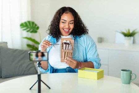 Photo for Happy black body positive blogger woman making makeup video tutorial for her blog, filming on smartphone and showing cosmetic brushes to phone camera - Royalty Free Image