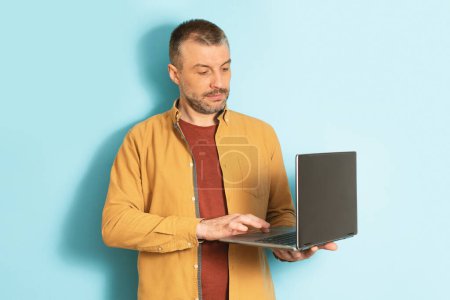 Photo for Smart businessman, employee working at computer. Serious mature man typing on laptop, standing on blue background, studio shot, free space - Royalty Free Image