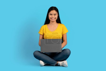Photo for Cheerful millennial korean woman student blogger in yellow t-shirt sit on floor with laptop, watch video, chatting in social networks, isolated on blue background. Device for work and study remote - Royalty Free Image