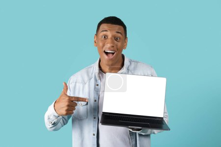 Téléchargez les photos : Choqué heureux excité jeune homme afro-américain avec bouche ouverte doigt pointeur à l'ordinateur portable avec écran blanc, isolé sur fond bleu. Étude, travail et affaires à distance, victoire en ligne, offre et annonce - en image libre de droit