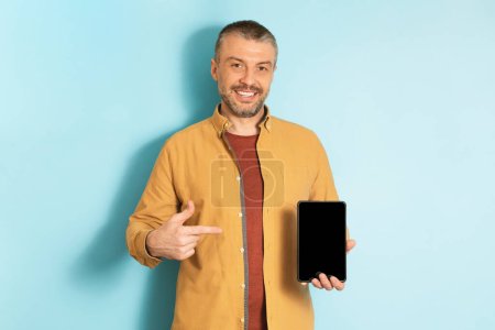Photo for Happy mature man demonstrating digital tablet and pointing at empty screen on blue studio background, mockup for website, app or advertisement design - Royalty Free Image