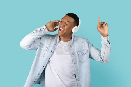 Photo for Cheerful young african american man in casual and wireless headphones sing song and enjoy music alone, isolated on blue background, studio, copy space. Audio app for fun and free time, rest and relax - Royalty Free Image