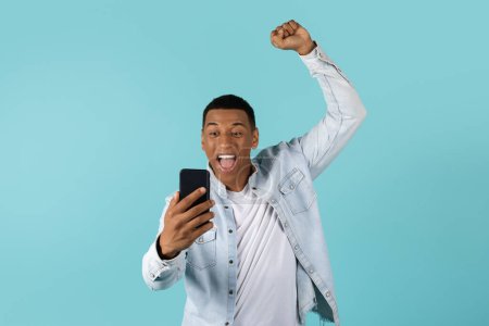 Téléchargez les photos : Heureux jeune homme afro-américain excité dans décontracté avec bouche ouverte regarder smartphone lève la main faisant geste de victoire et de succès, célébrer la victoire, isolé sur fond bleu, studio. Wow, bonne nouvelle - en image libre de droit