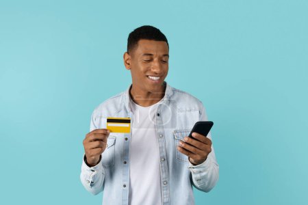 Foto de Sonriendo joven afroamericano masculino en casual mirando el teléfono inteligente, utiliza la tarjeta de crédito para ir de compras en línea, aislado en el fondo azul, estudio. Aplicación para banca financiera y venta, shopaholic remoto - Imagen libre de derechos
