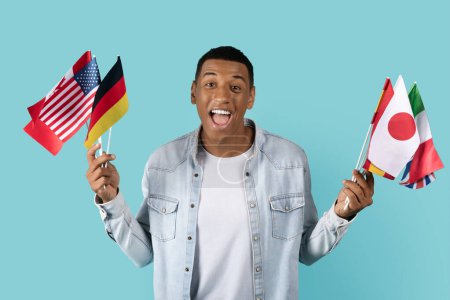 Photo for Glad young african american guy student in casual with open mouth shows many flags of different countries, isolated on blue background, studio. Learn language, tourism, education and study exchange - Royalty Free Image