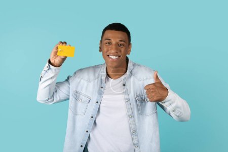Photo for Cheerful young african american guy in casual show credit card and thumb up, recommends for shopping online, isolated on blue background, studio. Advice of shopaholic, finance and huge sale, cashback - Royalty Free Image