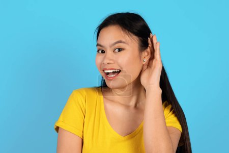 Portrait of happy inquisitive young korean lady in yellow t-shirt sticks out ear and listens news, looks at camera, isolated on blue background, studio. Lifestyle, gossip, ad and offer, human emotions