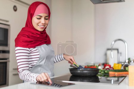 Photo for Happy middle eastern young woman wearing hijab enjoying cooking at home, checking online recipe or reading food blog on Internet, using digital tablet, side view, copy space - Royalty Free Image