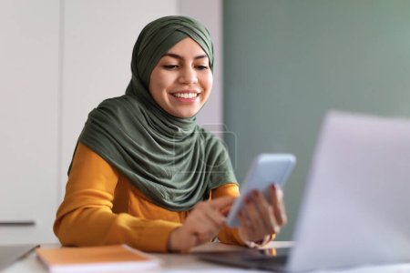 Téléchargez les photos : Femme musulmane souriante Dans Hijab Shopping en ligne avec Smartphone tout en étant assis au bureau au bureau, Happy Young Islamic Lady Browsing App sur téléphone mobile, Profiter de l'achat dans Internet, Gros plan - en image libre de droit
