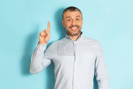 Photo for Eureka, gestures concept. Excited mature caucasian man pointing index finger up and smiling over blue studio background, having nice idea, copy space - Royalty Free Image
