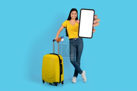 Téléchargez les photos : Joyeux joyeux blogueur étudiante coréenne millénaire avec valise, billets montrent smartphone avec écran blanc, isolé sur fond bleu, studio. App pour Voyage, vacances et tourisme, annonce, offre - en image libre de droit
