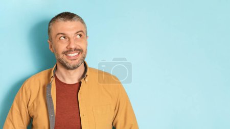 Téléchargez les photos : Portrait d'un homme d'âge moyen souriant posant sur fond bleu studio et regardant l'espace de copie, panorama. beau mâle mature montrant des émotions positives - en image libre de droit