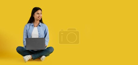 Photo for Cheerful cute millennial korean lady student in casual sit on floor, typing on computer, look at blank space, isolated on yellow background, studio. Device for study, work and business, ad and offer - Royalty Free Image