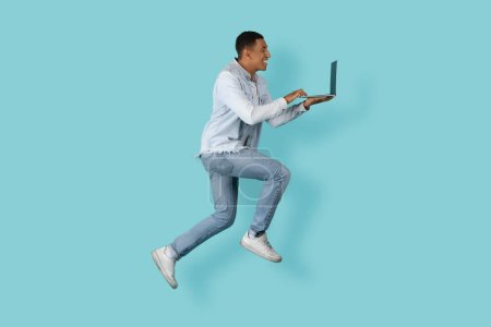 Photo for Happy busy young african american man student or manager typing on laptop, chatting, jumped, frozen in air isolated on blue background, studio. Technology for work, study, deadline and new normal - Royalty Free Image