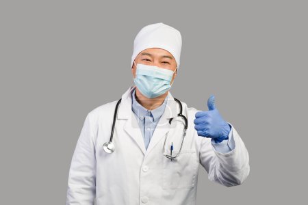 Photo for Smiling middle aged chinese male therapist in white coat, protective mask with stethoscope showing thumb up isolated on gray background. Doctor recommendation and advice, health care during covid-19 - Royalty Free Image
