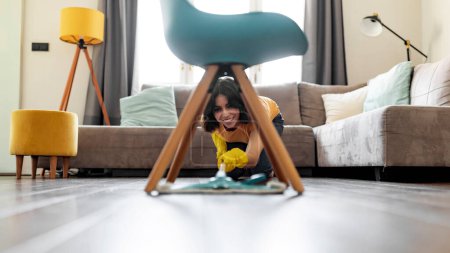 Téléchargez les photos : Souriant jeune femme arabe sol de nettoyage sous la chaise à la maison, joyeuse femme du Moyen-Orient en utilisant la serpillière tout en nettoyant le salon moderne, dame qui aime faire des corvées domestiques, espace libre - en image libre de droit
