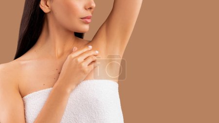 Photo for Cropped of long-haired brunette woman wrapped in towel with her arm up showing clean hairless armpit, panorama with copy space. Isolated on beige studio background. Epilation, hair removal concept - Royalty Free Image