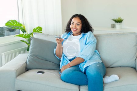 Foto de Feliz afroamericano más mujer de tamaño viendo la televisión, cambiando canales con mando a distancia, sentado en el sofá en casa. Programación televisiva y ocio de fin de semana - Imagen libre de derechos