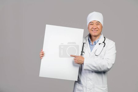 Foto de Terapeuta masculino chino maduro alegre en capa blanca con el estetoscopio sostiene la bandera con el espacio de la copia, aislado en fondo gris, estudio. Recomendación del médico, examen médico, atención médica, tratamiento - Imagen libre de derechos