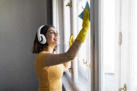 Téléchargez les photos : Souriant jeune femme arabe laver la fenêtre à la maison avec Rag, Enthousiaste Moyen-Orient féminin portant des écouteurs sans fil nettoyage maison, Écouter de la musique tout en faisant des tâches domestiques, Copier l'espace - en image libre de droit