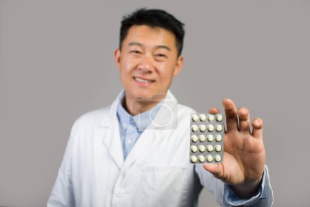 Foto de Sonriente terapeuta masculino chino de mediana edad en bata blanca mostrando píldoras, tratamiento médico recomendado, aislado sobre fondo gris, estudio, de cerca. Farmacia cuidado de la salud, consejo médico, oferta y anuncio - Imagen libre de derechos