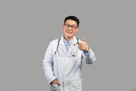 Photo for Cheerful middle aged japanese male therapist in white coat and glasses showing thumb up, advice medicine, isolated on gray background, studio. Doctor recommendation, health care and great services - Royalty Free Image