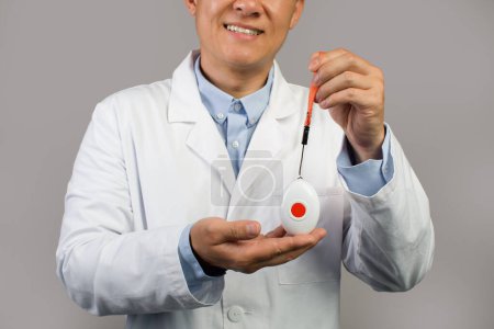 Foto de Sonriente terapeuta chico chino maduro en bata blanca muestran sos botón de alarma, aislado sobre fondo gris, recortado, de cerca. Ayuda, atención médica, atención médica y tratamiento para personas con discapacidad - Imagen libre de derechos