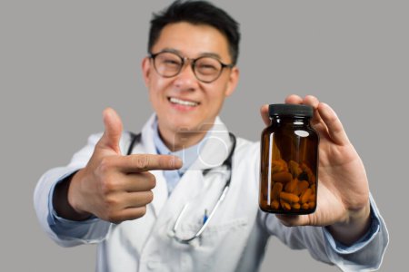 Téléchargez les photos : Un thérapeute chinois adulte souriant en manteau blanc et lunettes pointe du doigt le pot de médicaments, isolé sur fond gris. Conseils de médecin, choix pour le traitement, pilules de pharmacie pour les soins de santé - en image libre de droit
