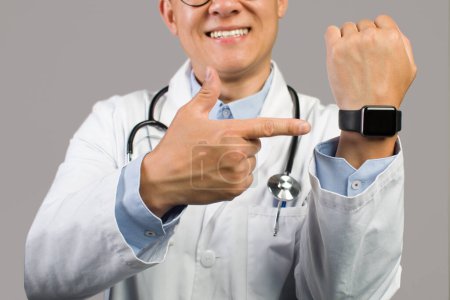 Téléchargez les photos : Heureux adulte asiatique homme médecin en manteau blanc pointant du doigt à la montre avec écran vide, isolé sur fond gris, studio, gros plan, recadré. Temps de travail et soins de santé, aide médicale et mode de vie - en image libre de droit