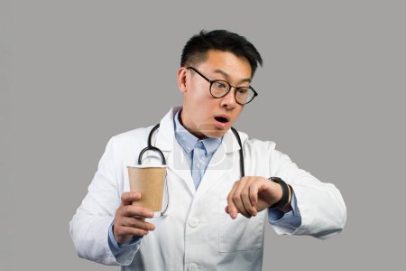 Photo for Shocked funny mature chinese male therapist in white coat, glasses with cup of coffee takeaway looks at watch, isolated on gray background, studio. Late for appointment and work and people emotions - Royalty Free Image