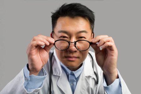 Photo for Serious suspects middle aged asian male therapist in white coat takes off glasses, looks at camera, isolated on gray background, close up, studio. Doctor checkup, treatment, health care examination - Royalty Free Image