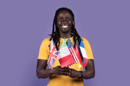 Photo for Happy handsome long-haired millennial african american guy in yellow t-shirt holding diverse flags of various countries and smiling over purple studio background. Education abroad, immigration concept - Royalty Free Image