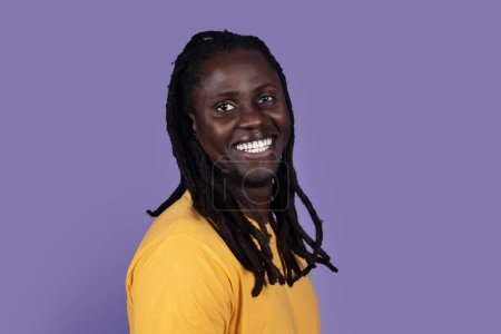 Téléchargez les photos : Gros plan portrait de heureux afro-américain millénial gars avec de longues tresses hipster souriant sur fond de studio violet, portant un t-shirt jaune, espace de copie. Noirs visages concept - en image libre de droit
