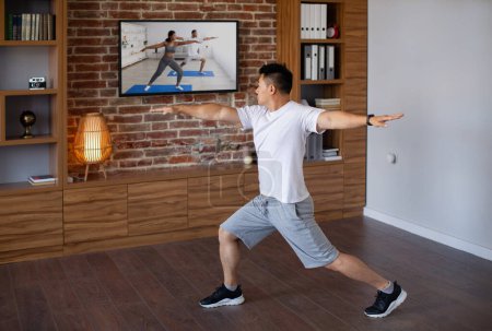 Photo for Domestic workout concept. Active asian mature man doing forward lunge stretch, watching fitness workout program on TV. Fitness and sport concept - Royalty Free Image