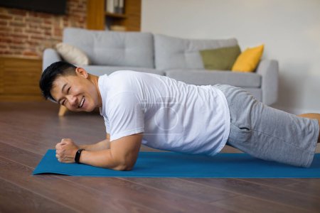 Foto de Feliz asiático maduro hombre haciendo fitness entrenamiento en deportes mat en casa, de pie en codo tablón y sonriendo en cámara. Emocionado hombre de mediana edad que lleva un estilo de vida saludable - Imagen libre de derechos