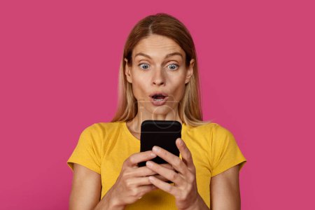 Photo for Glad surprised mature caucasian blonde female in yellow t-shirt with open mouth looks at smartphone, isolated on pink background, studio, close up. Amazing offer and ad, sale and app, win and news - Royalty Free Image