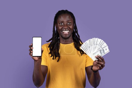 Foto de Apuesta en línea, comercio. Feliz hombre afroamericano joven de pelo largo en camiseta amarilla que muestra el teléfono celular moderno con pantalla blanca en blanco y montón de dólares en efectivo, fondo de estudio púrpura, maqueta - Imagen libre de derechos