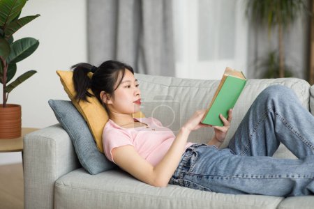 Foto de Señora coreana despreocupada acostada en el sofá en la sala de estar, lectura de libro mientras descansa en casa el fin de semana, espacio para copiar. Hobby, concepto de entretenimiento doméstico - Imagen libre de derechos